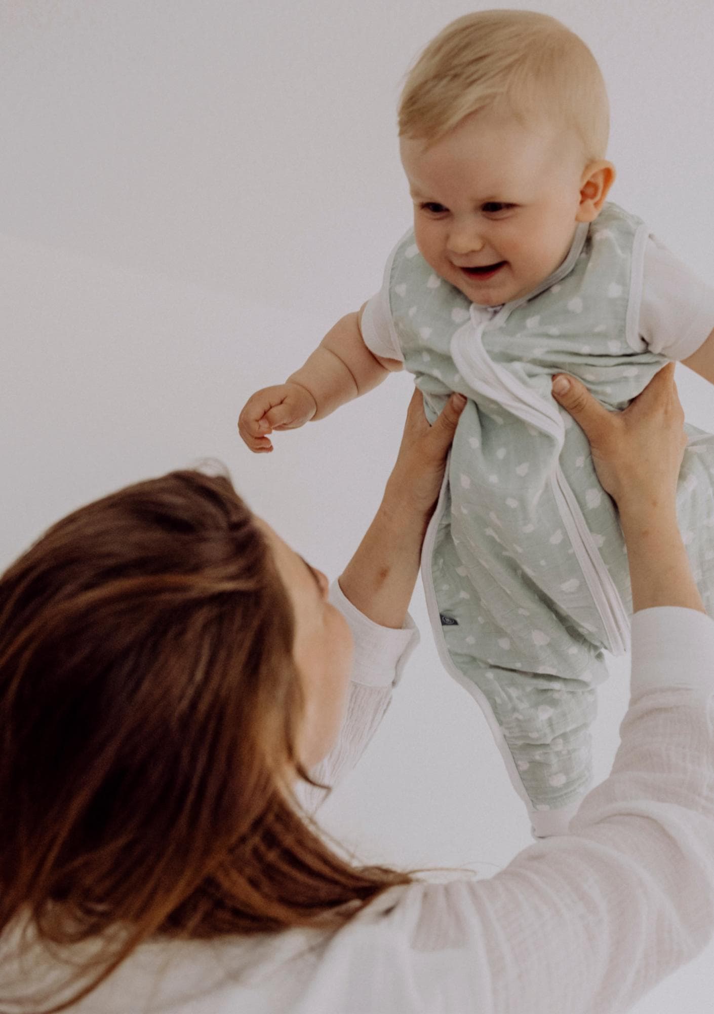 A woman with brown hair lifts a baby above her head. The baby wears a polka-dotted onesie with short sleeves. It's laughing.
