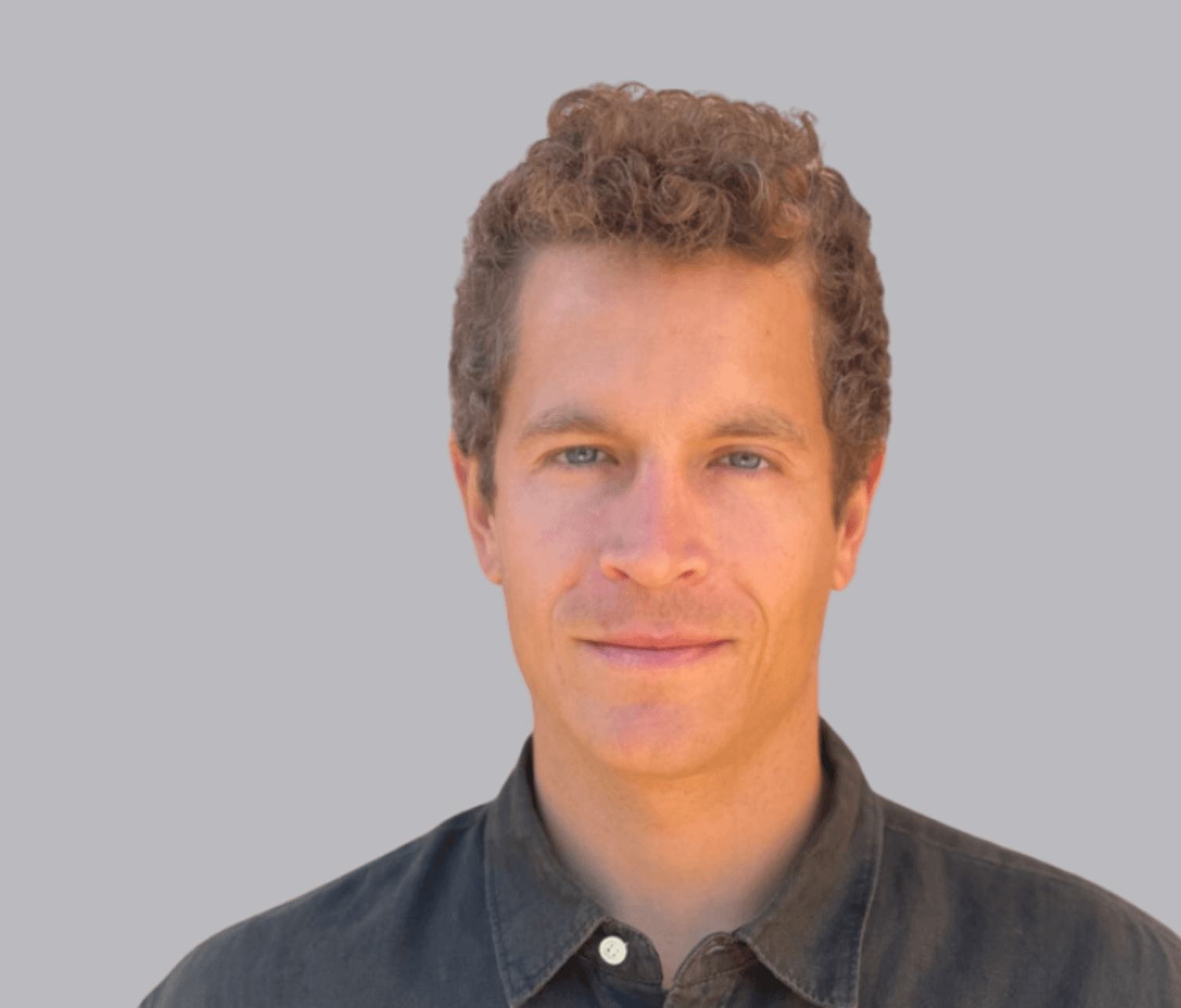 A middle-aged man looks at the camera. He smiles gently. He wears a black shirt and has short curly hair.