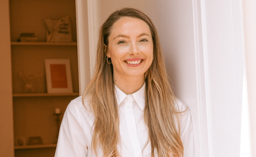 Woman with long blonde hair looks at the camera and smiles. She wears a white blouse and gold earrings.