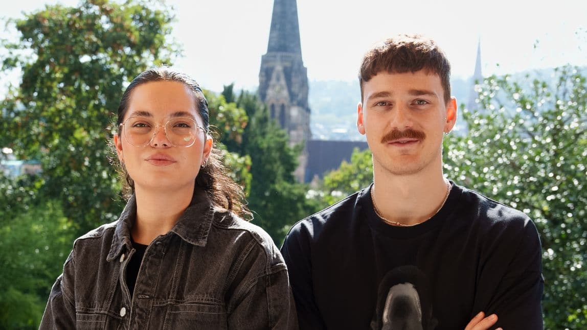 Two young people, a man and a woman, look at the camera. They stand outside, with bushes and a city skyline in the background. They look friendly.