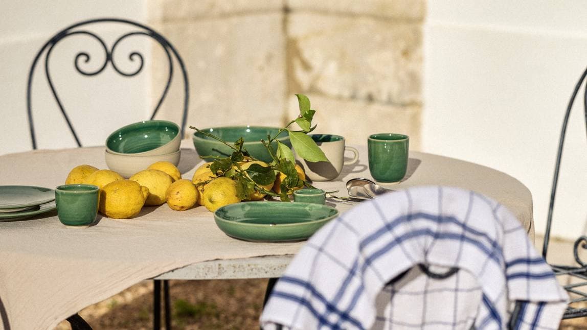 A set garden table with ornate black chairs around it. On the table, green ceramic dishes and lemons.