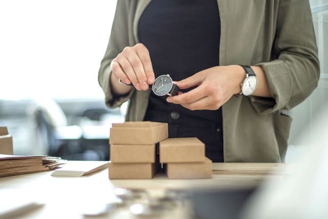 A woman holds a watch in her hand and has a watch on her wrist. She is packing watches.