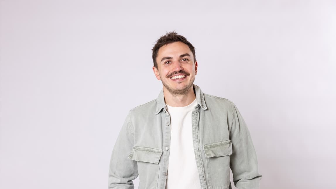 A man with short hair and a mustache looks into the camera laughing. He wears a white T-shirt and a green casual jacket over it.
