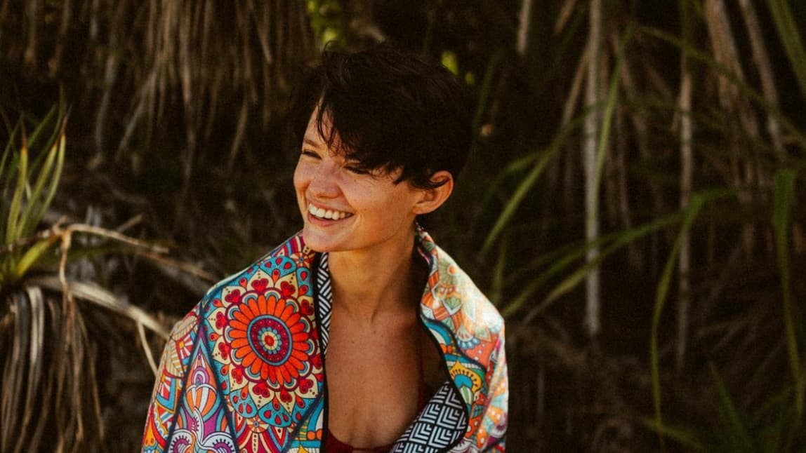 A woman with short black hair is at the beach. She looks to the side and laughs. She wears a colorful bikini and a colorful towel over it.